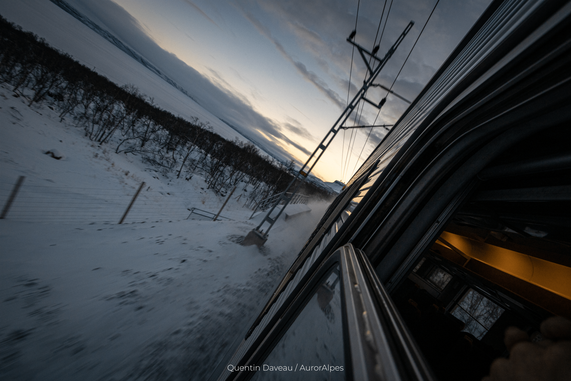 Vue depuis le train de nuit reliant Stockholm (Suède) à Narvik (Norvège). L'appareil photo est sorti en dehors du train, qui avance à vive allure. Au loin, le Soleil se lève sur un lac gelé.