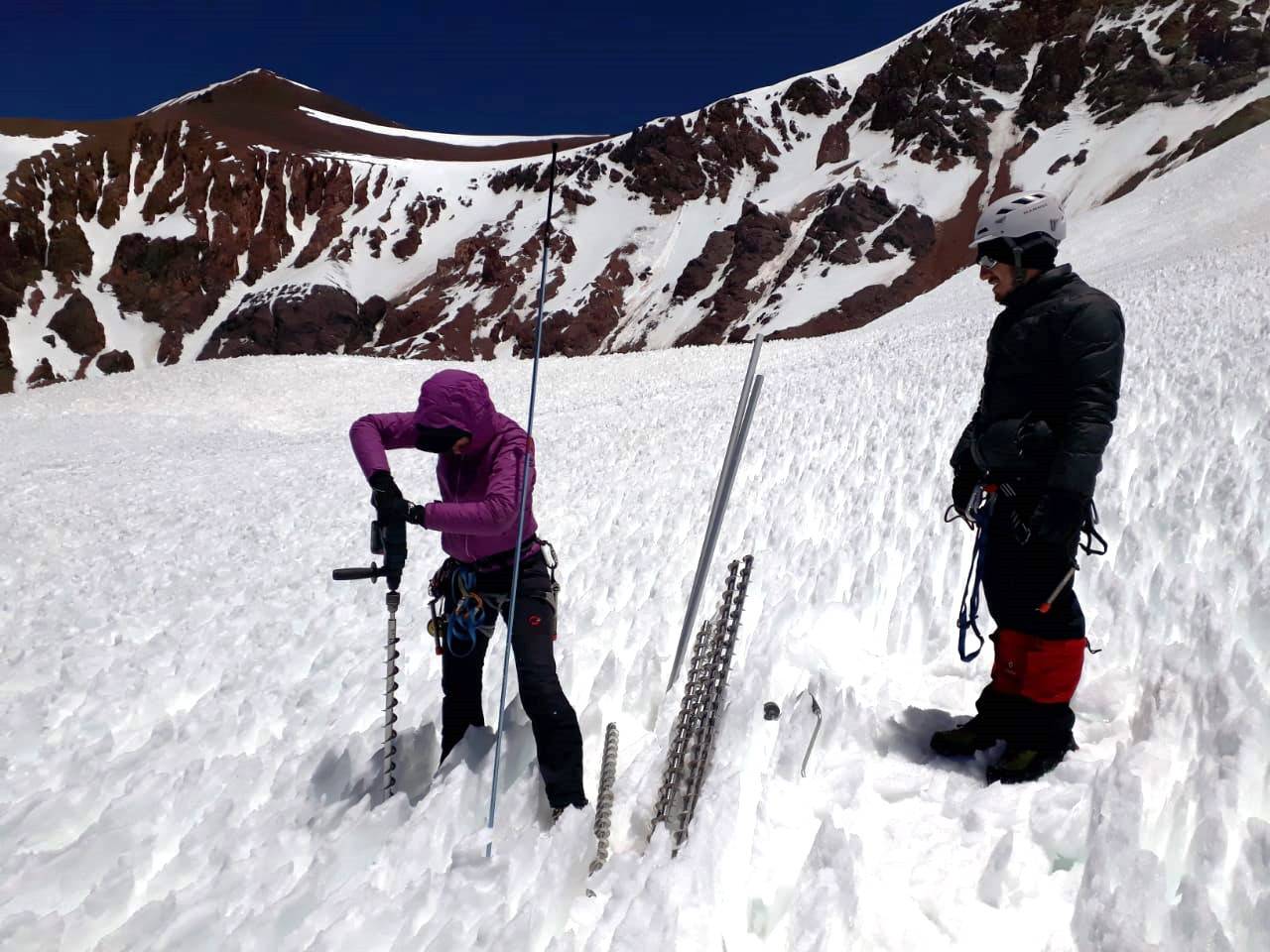 Photographie de Marion Réveillet et son collègue sur un glacier. Marion est courbée en train de planter une tige en fer dans la glace. Son collègue la regarde.